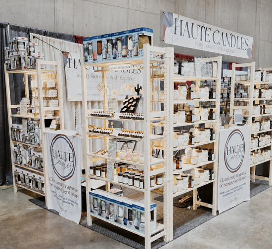 A booth display for Haute Candles at Astoria Crab and Wine Festival featuring multiple light wood shelving units stocked with various scented candles, wax melts, and packaged fragrance products, set against a concrete wall background.